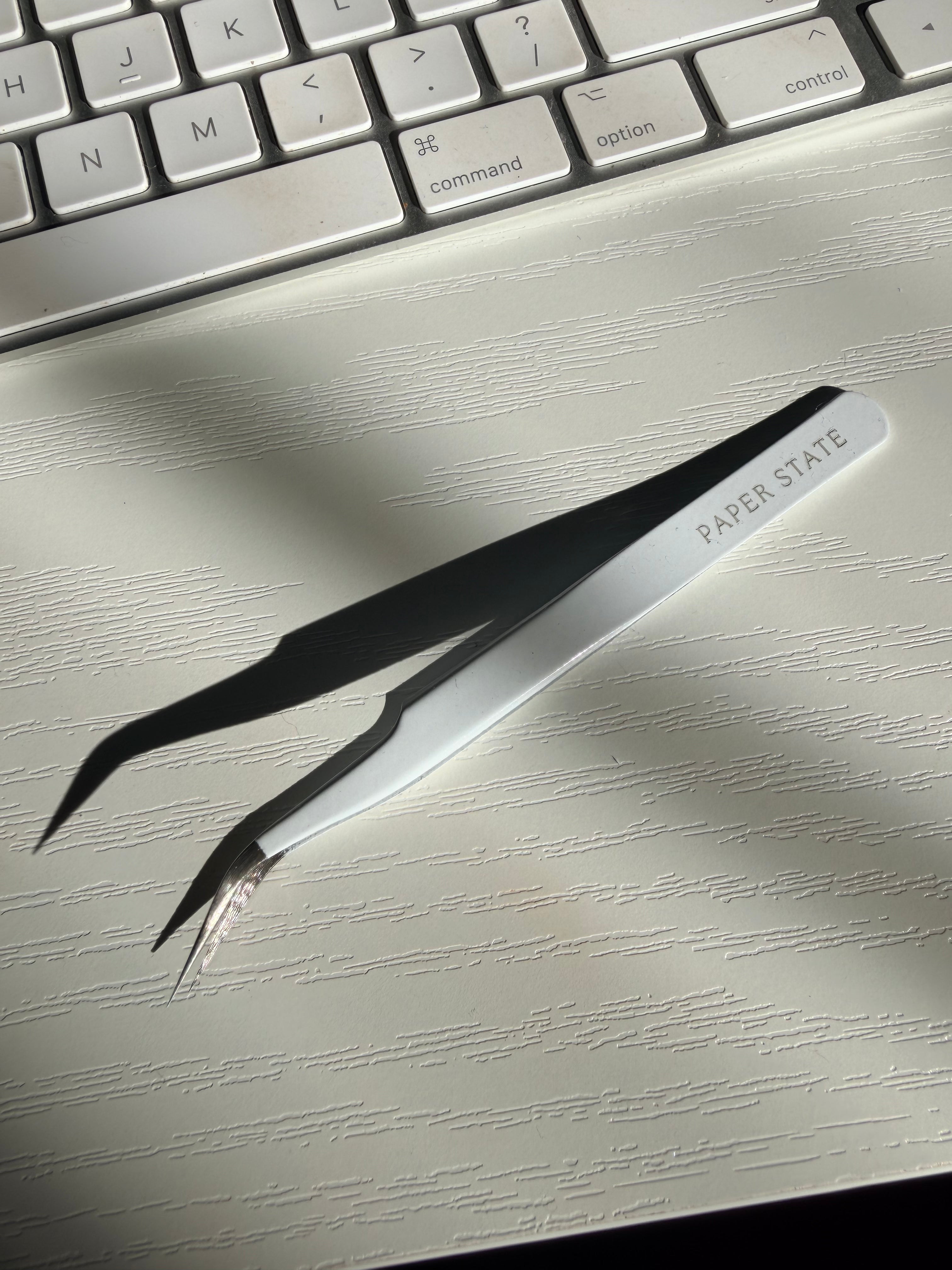 White and black precision tweezers with 'PAPER STATE' printed on the handle, resting on a light textured surface with a keyboard in the background.